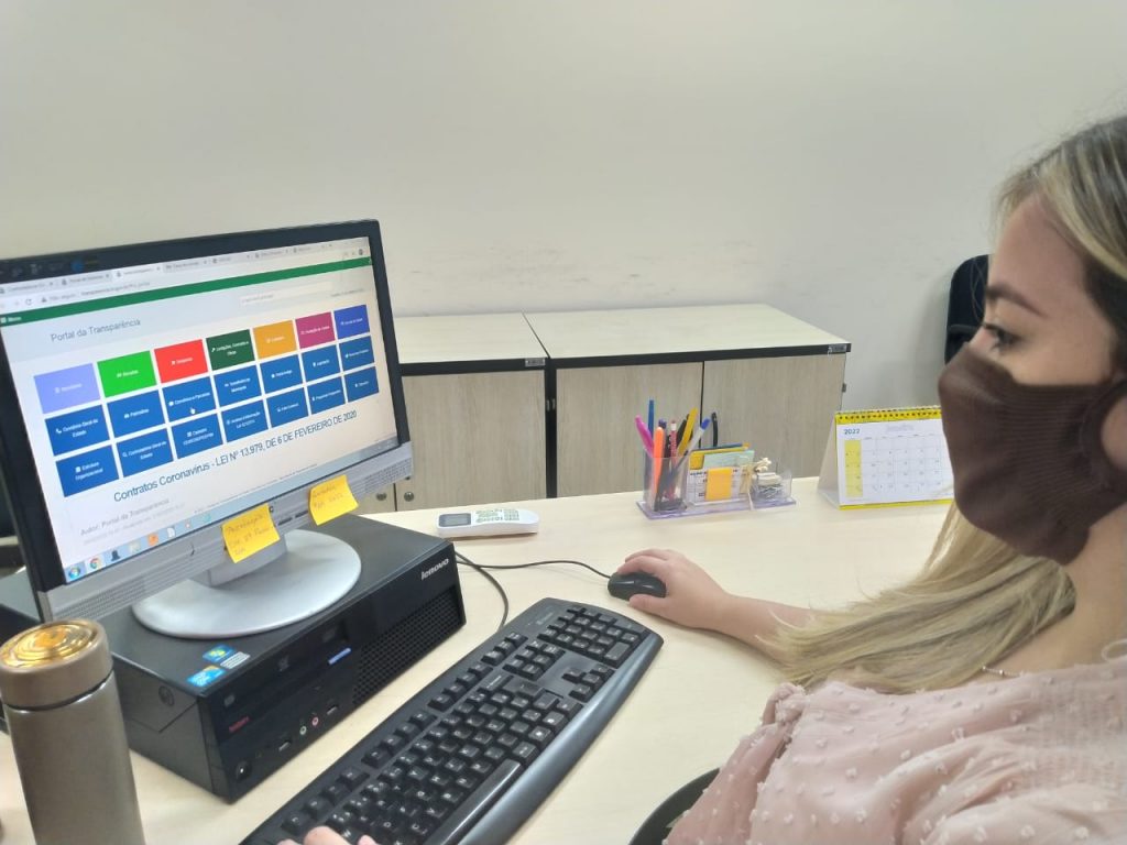 Portal Jaciara Barros – Uma mulher com máscara marrom utiliza um computador no escritório para acessar o Portal da Transparência Tocantins. – Portal Jaciara Barros