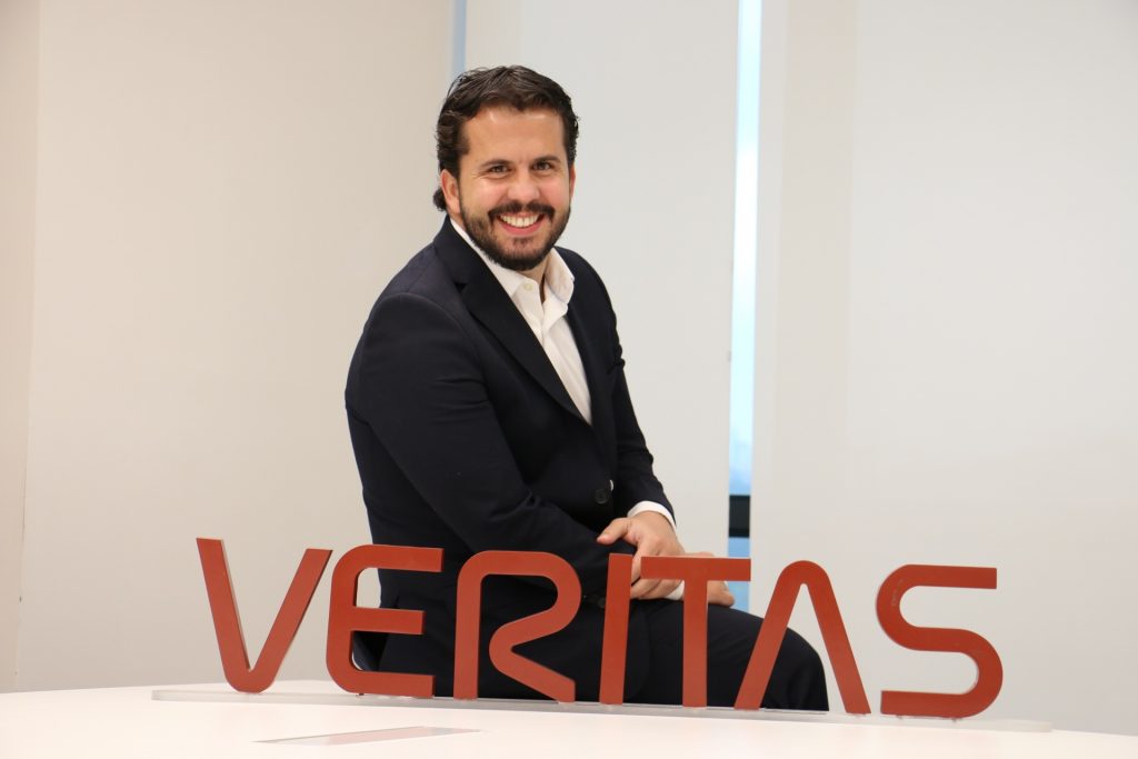 Portal Jaciara Barros – Um CEO de terno escuro está sentado em uma mesa, sorrindo, com uma placa vermelha da VERITAS posicionada à sua frente. – Portal Jaciara Barros