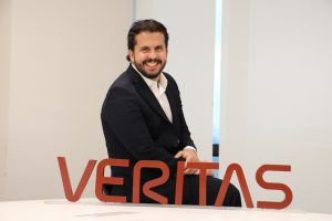 Portal Jaciara Barros – Um CEO de terno escuro está sentado em uma mesa, sorrindo, com uma placa vermelha da VERITAS posicionada à sua frente. – Portal Jaciara Barros