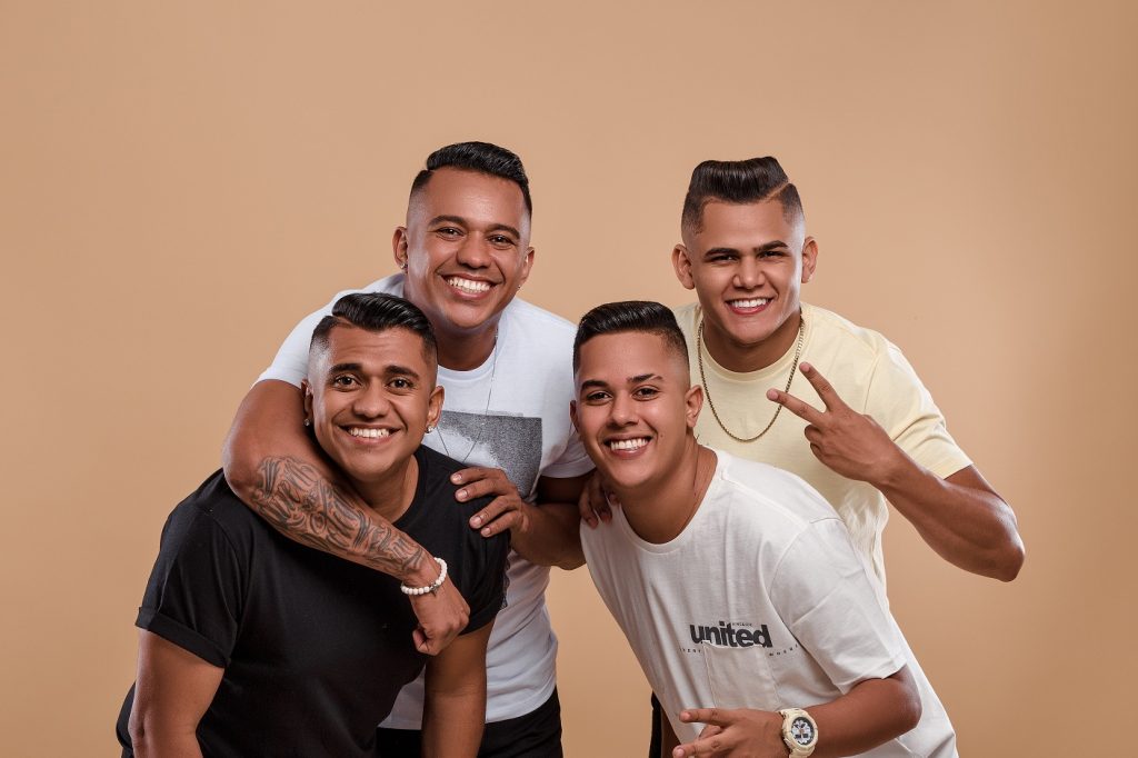 Portal Jaciara Barros – Quatro jovens posam sorrindo, um faz sinal de paz, em clima de pagode durante gravação de DVD com fundo bege. – Portal Jaciara Barros