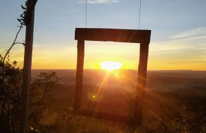 Portal Jaciara Barros – Um quadro de madeira ao ar livre enquadra o pôr do sol sobre as colinas e a variedade de árvores de Serras Gerais. – Portal Jaciara Barros
