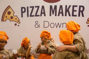 Portal Jaciara Barros – Crianças com síndrome de Down, usando chapéus de chef laranja, posam em frente à placa do Pizza Maker & Down. – Portal Jaciara Barros
