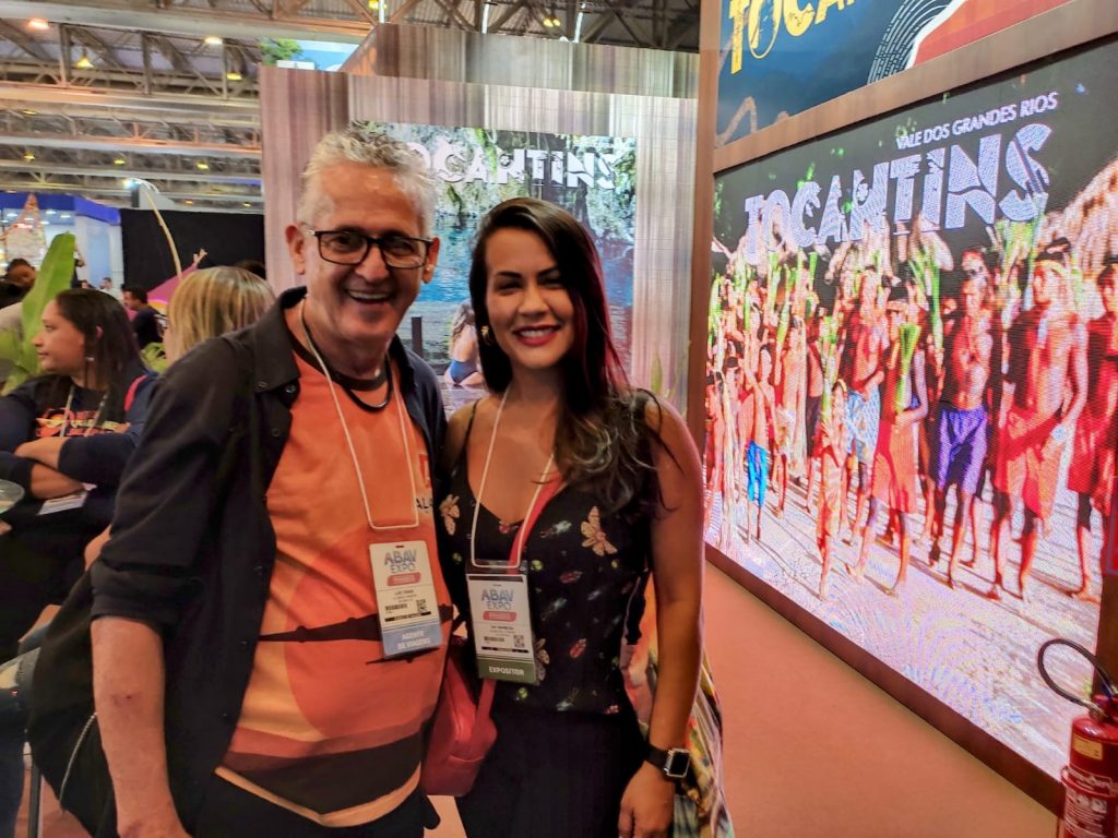 Portal Jaciara Barros – Day Barbosa e Luiz César posam sorrindo diante de um estande do Tocantins durante evento ou convenção em local fechado. – Portal Jaciara Barros