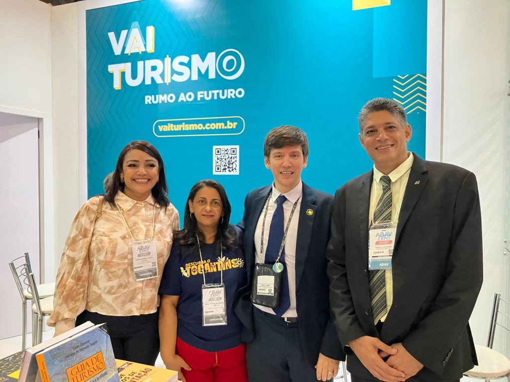 Portal Jaciara Barros – Quatro pessoas estão sorrindo diante do estande azul e amarelo da Vai Turismo na ABAV Expo. – Portal Jaciara Barros