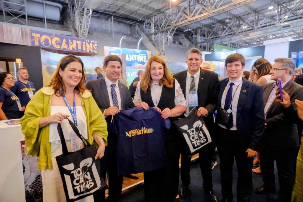 Portal Jaciara Barros – Grupo posa sorridente com bolsas e camisa do Tocantins no estande do estado durante evento realizado em ambiente fechado. – Portal Jaciara Barros