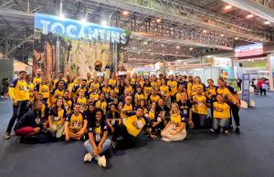 Portal Jaciara Barros – Integrantes da Caravana Tocantins, com camisas amarelas e azuis, posam para foto oficial em evento interno com painel do Tocantins. – Portal Jaciara Barros