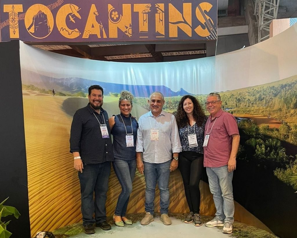 Portal Jaciara Barros – Cinco pessoas posam sorrindo diante de um painel de Tocantins na ABAV Expo, com paisagens e logomarca da Febtur ao fundo. – Portal Jaciara Barros