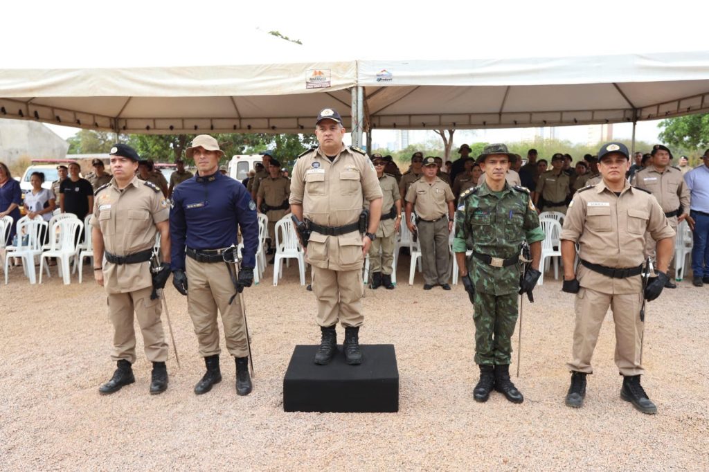 Portal Jaciara Barros – Cinco policiais militares estão em frente a uma tenda durante troca de comando, com público sentado ao fundo. – Portal Jaciara Barros