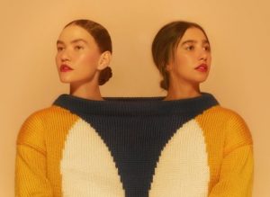 Portal Jaciara Barros – Duas mulheres usam juntas um suéter estampado, em referência à turnê Cor de Anavitória, sobre fundo pêssego. – Portal Jaciara Barros