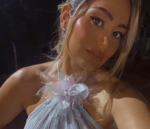 Portal Jaciara Barros – A médica Analú Barros registra selfie em close, usando vestido lilás com detalhes florais, em fundo escuro. – Portal Jaciara Barros