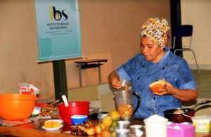 Portal Jaciara Barros – Mulher de lenço prepara comida ao lado de placa do Instituto Brasil Sustentável, que incentiva vivência e sustentabilidade. – Portal Jaciara Barros