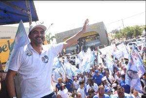 Portal Jaciara Barros – O governador Wanderlei Barbosa acena para o público com bandeiras durante caminhada histórica em Araguaína, sob sol forte. – Portal Jaciara Barros