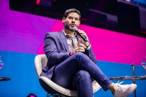 Alex Monteiro sentado em uma cadeira branca no Fire Festival 2022, iluminado por luzes de palco rosa e azuis. – Portal Jaciara Barros