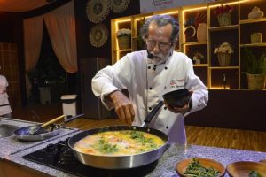 Portal Jaciara Barros – Chef renomado e experiente, de uniforme branco, adiciona ervas a uma panela grande de sopa em cozinha moderna. – Portal Jaciara Barros