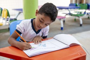 Portal Jaciara Barros – Um menino preenche um livro de atividades em uma carteira laranja na sala de aula do Cmei Sítio do Pica Pau Amarelo. – Portal Jaciara Barros