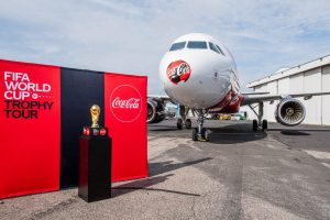 Portal Jaciara Barros – A Taça da Copa do Mundo exibe ao lado de banners da Coca-Cola e de um avião do Tour da Taça com o logotipo oficial. – Portal Jaciara Barros