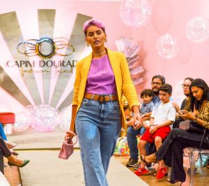 Portal Jaciara Barros – Modelo de cabelo rosa desfila com roupas coloridas em evento de moda, observado pelo público sentado dos dois lados da passarela. – Portal Jaciara Barros