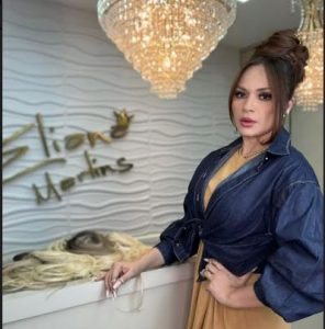 Portal Jaciara Barros – Durante o Goiás Fashion Week, uma mulher de vestido marrom e jaqueta jeans posa ao lado de perucas sob lustres, próxima à placa Eliona Merlins. – Portal Jaciara Barros