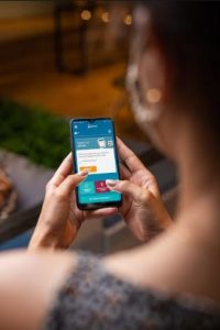 Portal Jaciara Barros – O usuário utiliza o aplicativo financeiro no smartphone em casa, com acesso digital e tela do aparelho visível. – Portal Jaciara Barros