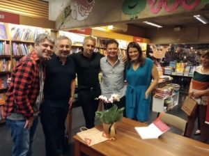 Portal Jaciara Barros – Cinco adultos posam sorrindo atrás de uma mesa com planta em evento da Expedição Antártida, realizado em livraria iluminada. – Portal Jaciara Barros