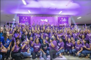 Portal Jaciara Barros – Um grupo de mulheres certificadas, vestindo camisetas roxas, posa em ambiente interno segurando cilindros roxos na vertical. – Portal Jaciara Barros