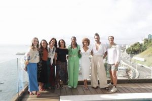 Portal Jaciara Barros – Oito pessoas posam lado a lado em uma varanda com vista para o mar e a estrada litorânea. – Portal Jaciara Barros