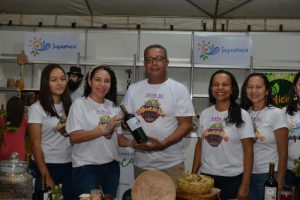 Portal Jaciara Barros – Cinco pessoas com camisetas do evento posam atrás de uma mesa da Festa da Colheita da Jabuticaba, com comida e vinho. – Portal Jaciara Barros