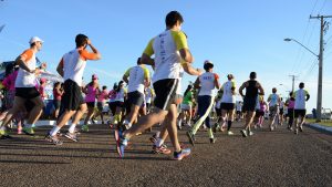 Portal Jaciara Barros – Atletas com uniformes esportivos e números participam da Corrida Hospital Jorge Saad sob sol em estrada asfaltada. – Portal Jaciara Barros