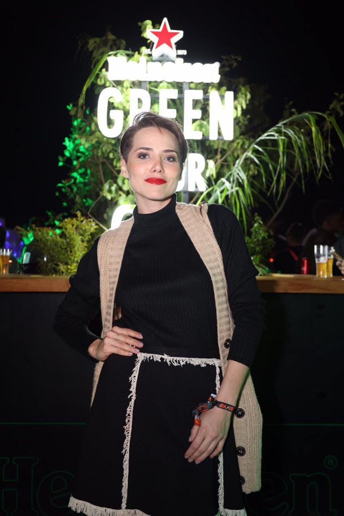 Portal Jaciara Barros – Uma pessoa, vestindo roupa preta e colete bege, posa diante do letreiro Heineken Lounge no Rock in Rio. – Portal Jaciara Barros
