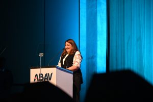 Portal Jaciara Barros – Durante a ABAV Expo, uma mulher discursa no palco iluminado de azul, destacando culturas como o frevo e o maracatu. – Portal Jaciara Barros