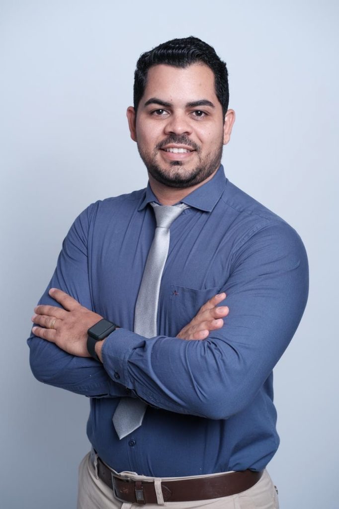 Portal Jaciara Barros – O médico Miguel Cláudio Silva, de cabelos escuros e barba, sorri para a câmera usando camisa azul e gravata prateada. – Portal Jaciara Barros