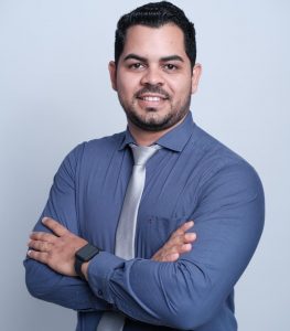 Portal Jaciara Barros – O médico Miguel Cláudio Silva, de cabelos escuros e barba, sorri para a câmera usando camisa azul e gravata prateada. – Portal Jaciara Barros