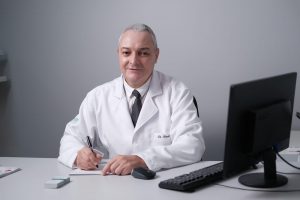 Portal Jaciara Barros – O oftalmologista Renato de Carvalho Costa, de branco, escreve à mesa cercado por monitor e materiais médicos. – Portal Jaciara Barros