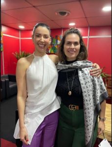 Portal Jaciara Barros – Duas mulheres posam sorrindo em um ambiente interno: uma veste blusa branca e calça roxa; a outra, xale e blusa escura. – Portal Jaciara Barros