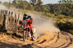 Portal Jaciara Barros – Piloto de motocross com roupa vermelha e capacete verde acelera nas rotas inéditas do Sertões BRB, levantando poeira perto de uma cerca. – Portal Jaciara Barros