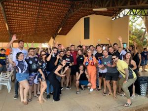 Portal Jaciara Barros – Um grande grupo de jovens posa sob um pavilhão, sorrindo e com as mãos erguidas para uma foto coletiva. – Portal Jaciara Barros