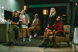 Portal Jaciara Barros – Cinco mulheres comemoram o movimento Heroínas do Game em um estúdio com espelhos de camarim, araras e luzes brilhantes. – Portal Jaciara Barros