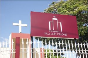 Portal Jaciara Barros – Cerca branca e portão diante da placa vinho da Paróquia São Luís Orione, local do animado Orione Fest, com árvore e céu ao fundo. – Portal Jaciara Barros