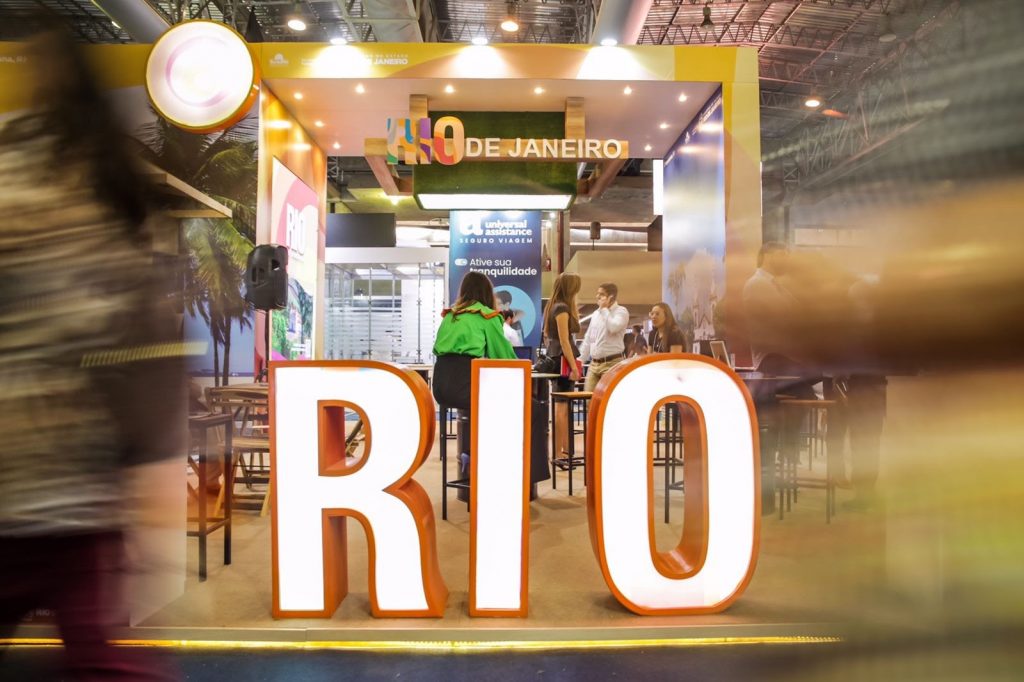Portal Jaciara Barros – Letreiro iluminado com a palavra RIO marca o estande Destino Anfitrião na ABAV Expo 2023, com público circulando. – Portal Jaciara Barros