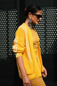 Portal Jaciara Barros – Uma mulher com cardigã amarelo Gucci e vestido estampado passa por uma parede preta perfurada em Milão. – Portal Jaciara Barros