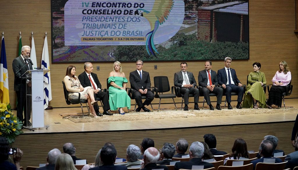 Portal Jaciara Barros – Em Palmas, um homem discursa no púlpito enquanto oito pessoas estão sentadas ao lado de uma faixa do Governo do Tocantins. – Portal Jaciara Barros