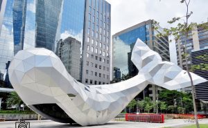 Portal Jaciara Barros – Escultura geométrica de cauda de baleia ocupa praça urbana, cercada pelo Edificio B32 e prédios modernos de vidro. – Portal Jaciara Barros
