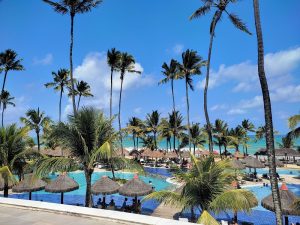 Portal Jaciara Barros – A piscina do resort em Porto exibe palmeiras, guarda-sóis de palha, banhistas e vista para o oceano ao fundo. – Portal Jaciara Barros
