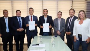 Portal Jaciara Barros – Luciano Casaroti e outro homem exibem documentos e sorriem diante de uma mesa de vidro, acompanhados por mais cinco pessoas. – Portal Jaciara Barros