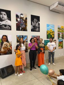 Portal Jaciara Barros – Cinco adultos e uma criança posam na Galeria AMAN durante exposição de jovens artistas tocantinenses e obras vibrantes. – Portal Jaciara Barros