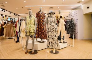 Portal Jaciara Barros – Quatro manequins exibem vestidos estampados da coleção C&A em loja bem iluminada, com clientes ao fundo. – Portal Jaciara Barros