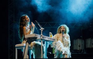 Portal Jaciara Barros – Anavitória se apresenta no Capim Dourado Shopping usando trajes brancos; uma toca teclado. Ambas são iluminadas por luz azul. – Portal Jaciara Barros