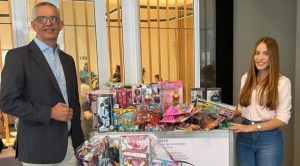 Portal Jaciara Barros – Um homem e uma mulher estão ao lado de uma mesa com brinquedos embalados para doação em um prédio comercial moderno. – Portal Jaciara Barros