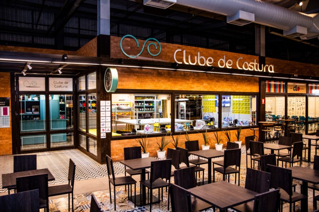 Portal Jaciara Barros – A fachada do Clube de Costura exibe mesas e cadeiras na área externa, com máquinas de costura visíveis pelas janelas. – Portal Jaciara Barros