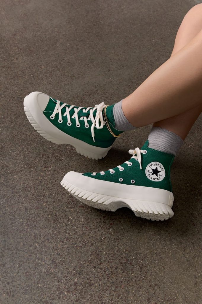 Portal Jaciara Barros – A pessoa usa tênis verdes Converse Coleção Lugged 2.0 de sola branca grossa e meia cinza, com as pernas cruzadas no piso manchado. – Portal Jaciara Barros